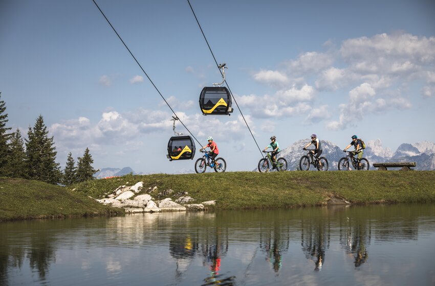 Four mountain bikers ride by a pond below two Planai gondolas, with mountains and forest landscape in the background | © Planai-Hochwurzen-Bahnen GesmbH