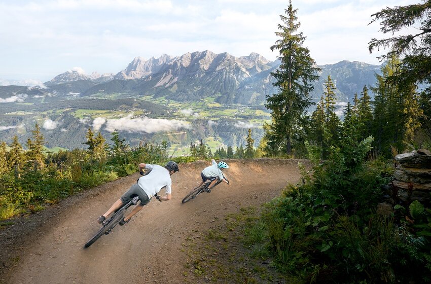 Two mountain bikers on a curved trail overlooking green valleys, forests and the Dachstein mountain range in the background | © Dietmar Koerbler