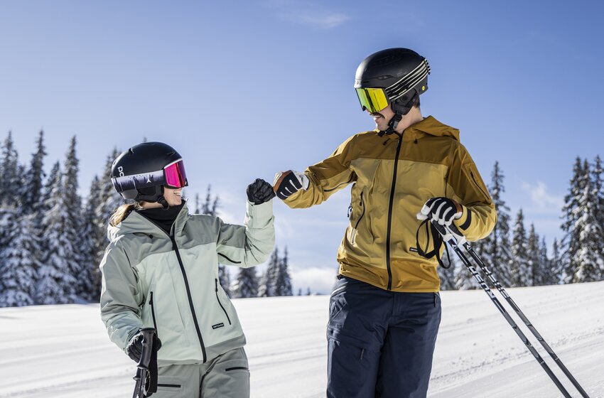 Two skiers wearing helmets and holding ski poles greet each other on a snowy slope with snow-covered fir trees. | © Mirja Geh