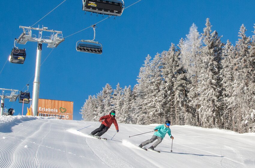 Two skiers enjoy a sunny descent below the Rittisberg Erlebnisbahn, with snowy forest beside the slope. | © Rittis-Lift Engelhardt GmbH & Co KG
