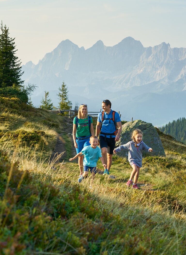 Family with two kids hiking happily on an alpine trail with mountain views and a distant mountain hut. | © SCHLADMING-DACHSTEIN Tourismusmarketing GmbH