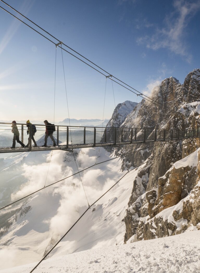 Four people cross the snowy Dachstein suspension bridge in sunshine with mountain views and drifting mist below. | © SCHLADMING-DACHSTEIN Tourismusmarketing GmbH