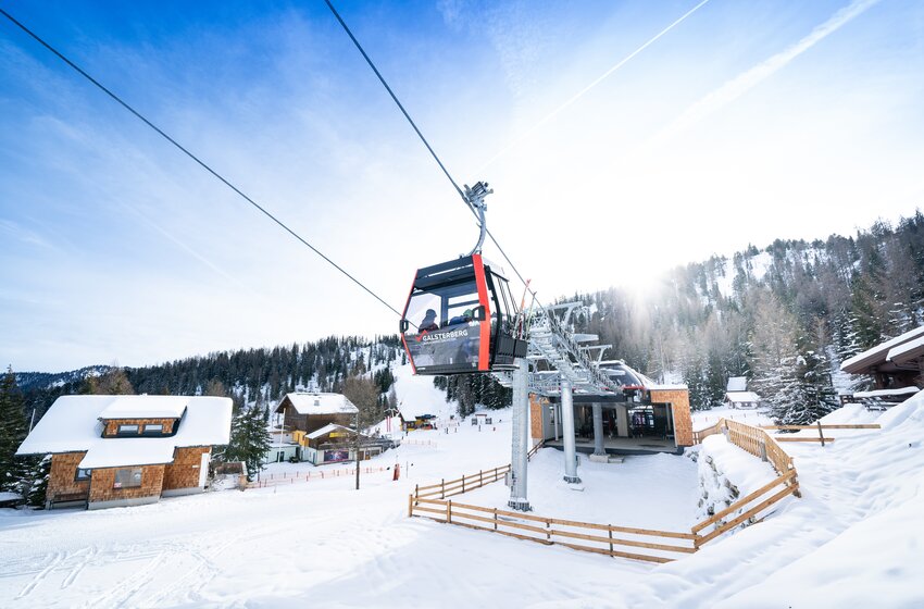 Red Galsterberg gondola near valley station in sunshine, surrounded by snowy cabins and forested mountain slopes. | © Galsterbergalm Bahnen GmbH & Co KG 