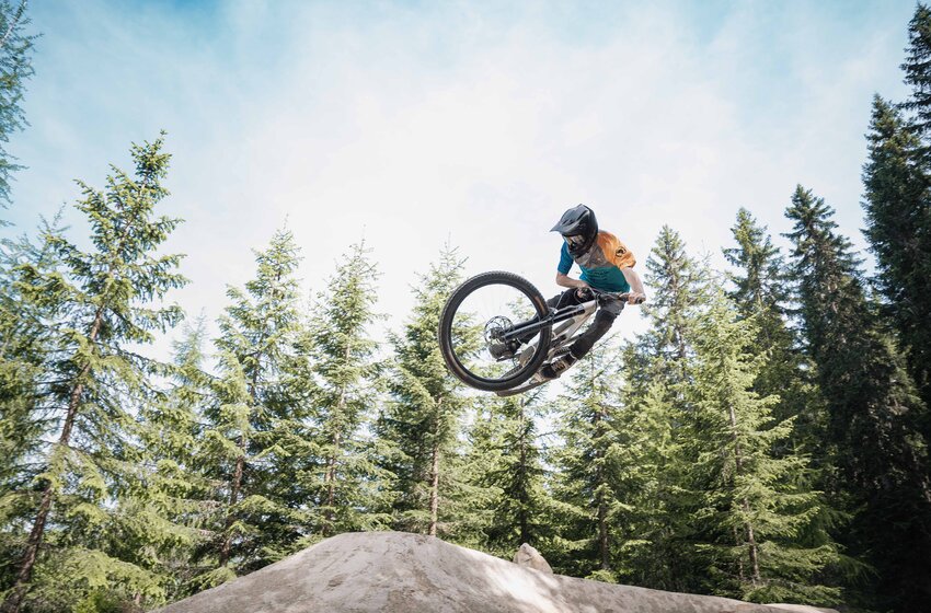 Mountain biker with helmet jumps on Reiteralm over dirt mound, tall evergreen trees in the background. | © Reiteralm Bergbahnen GmbH & Co.KG