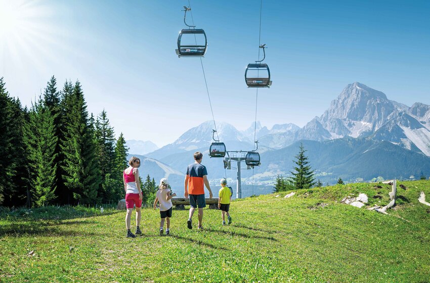 Familie wandert bei Sonnenschein durch grüne Almwiesen mit Gondelbahn und Bergpanorama im Hintergrund. | © Ski amadé GmbH