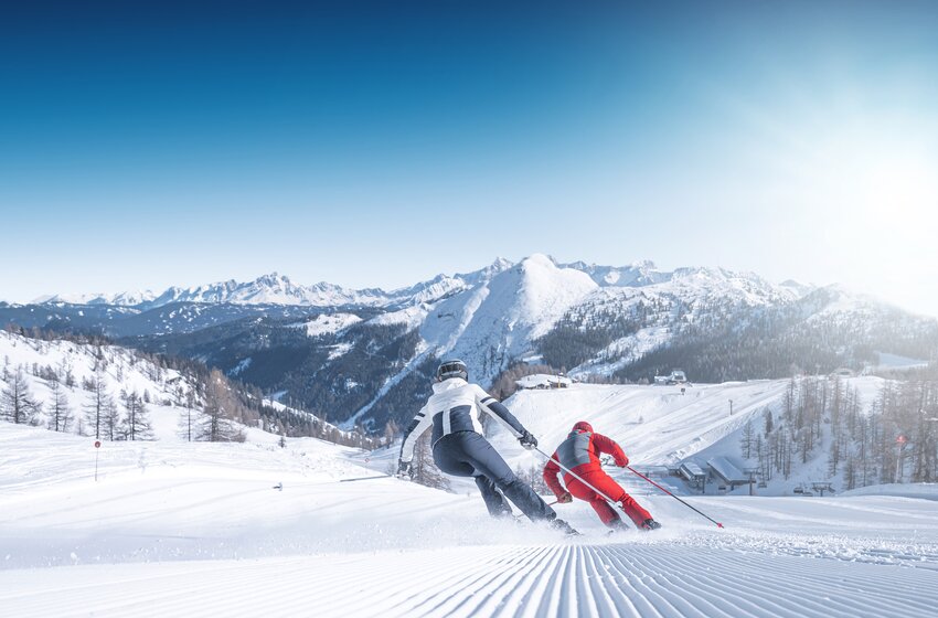 Zwei Skifahrer fahren bei Sonnenschein auf frisch präparierter Piste mit Panoramablick durch die verschneiten Berge von Ski amadé. | © Ski amadé GmbH